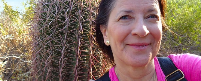 Woman wearing a pink shirt taking a selfie outdoors, with a large cactus behind her and desert vegetation in bright daylight.
