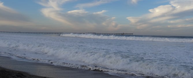 Beach scene with blue sky and wispy clouds, a long pier extending into the ocean, and waves rolling onto the sandy shore.