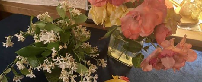 Colorful arrangement of pink and yellow roses in a glass vase with white blossoms nearby, and a lemon on a dark table in the foreground.