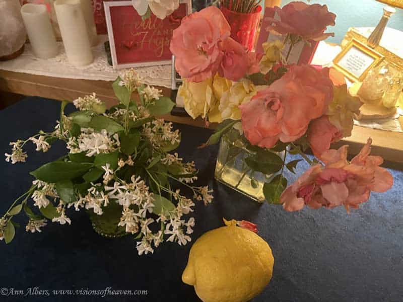 Colorful arrangement of pink and yellow roses in a glass vase with white blossoms nearby, and a lemon on a dark table in the foreground.